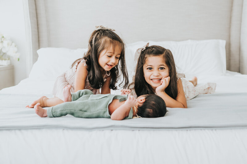 Two little girls sit on bed smiling with newborn baby brother during in home photography session