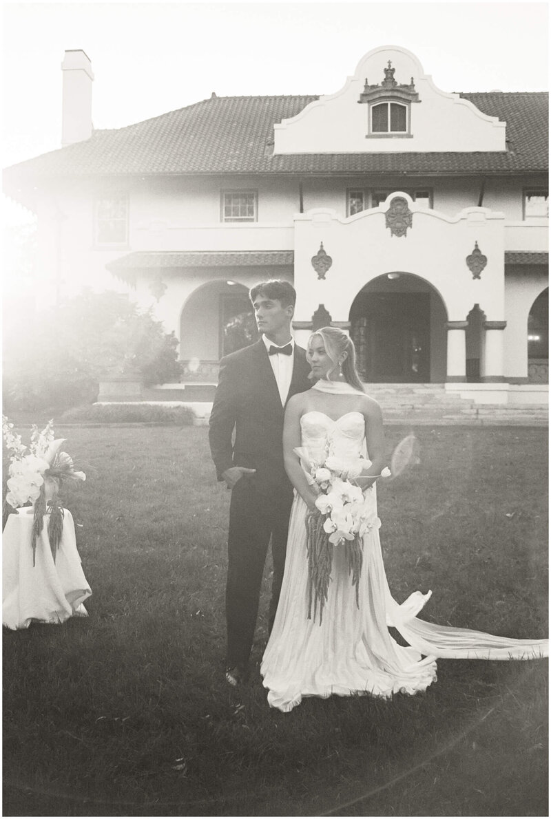 A bride leans into her groom as they look over their shoulders into the distance in the front lawn from a film wedding photographer