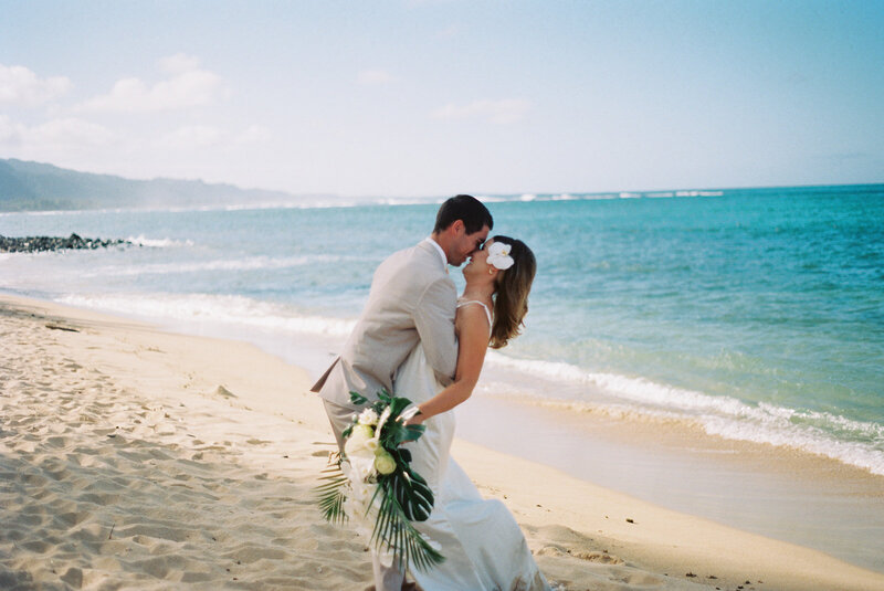 Newlyweds embrace face to face and lean back together in front of Hawaii beach