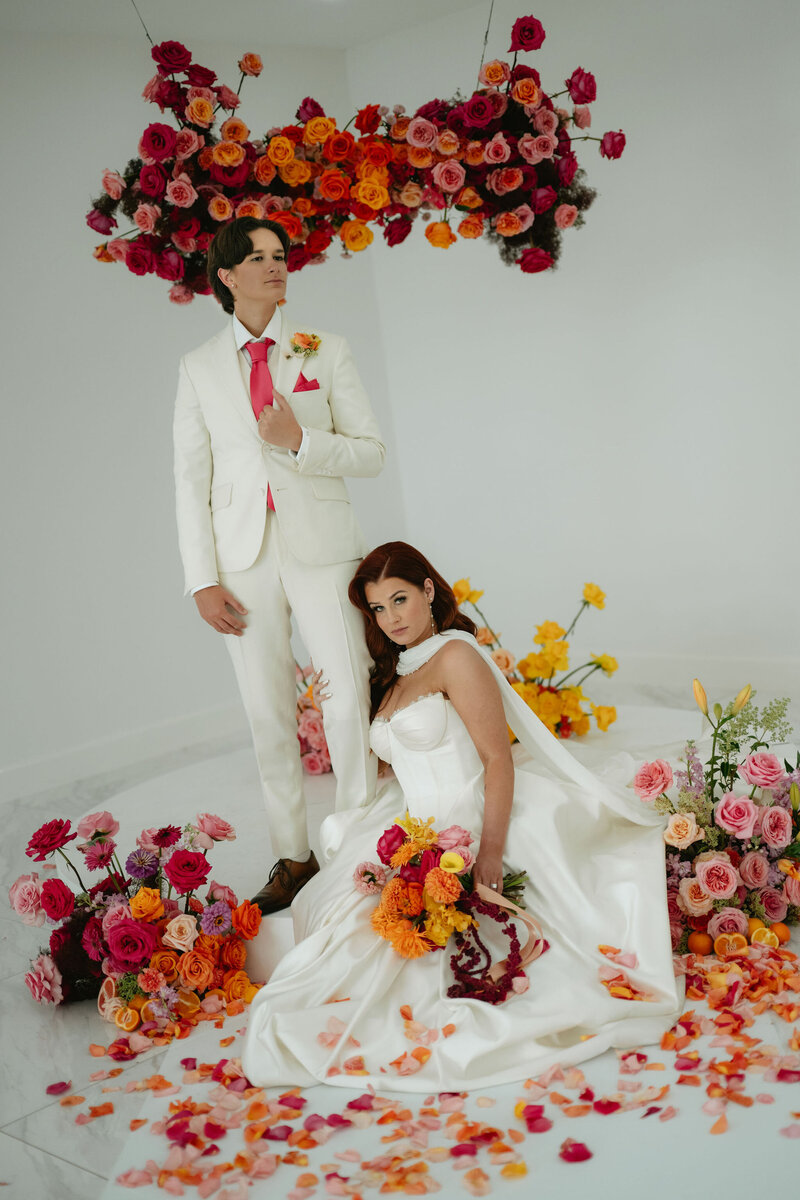 A couple in a white room with vibrant pink, orange, and yellow flowers. The bride in a white suit stands confidently, while the other bride in a white gown sits gracefully, holding a bouquet.