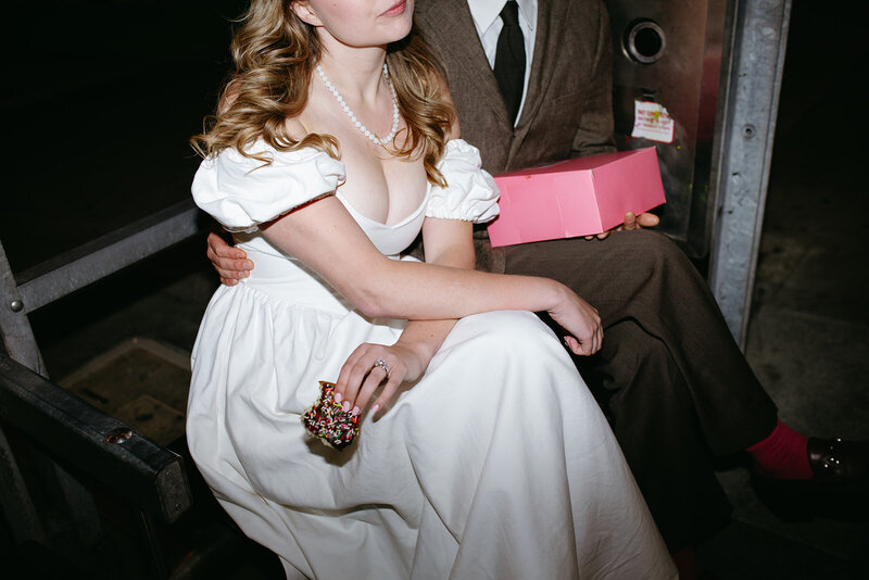 A bride and groom sit at a bus stop in San Francisco with a box of donuts. 