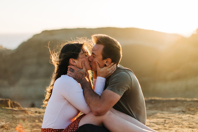 couple shares a passionate kiss sitting on the beach in pacific city oregon at golden hour