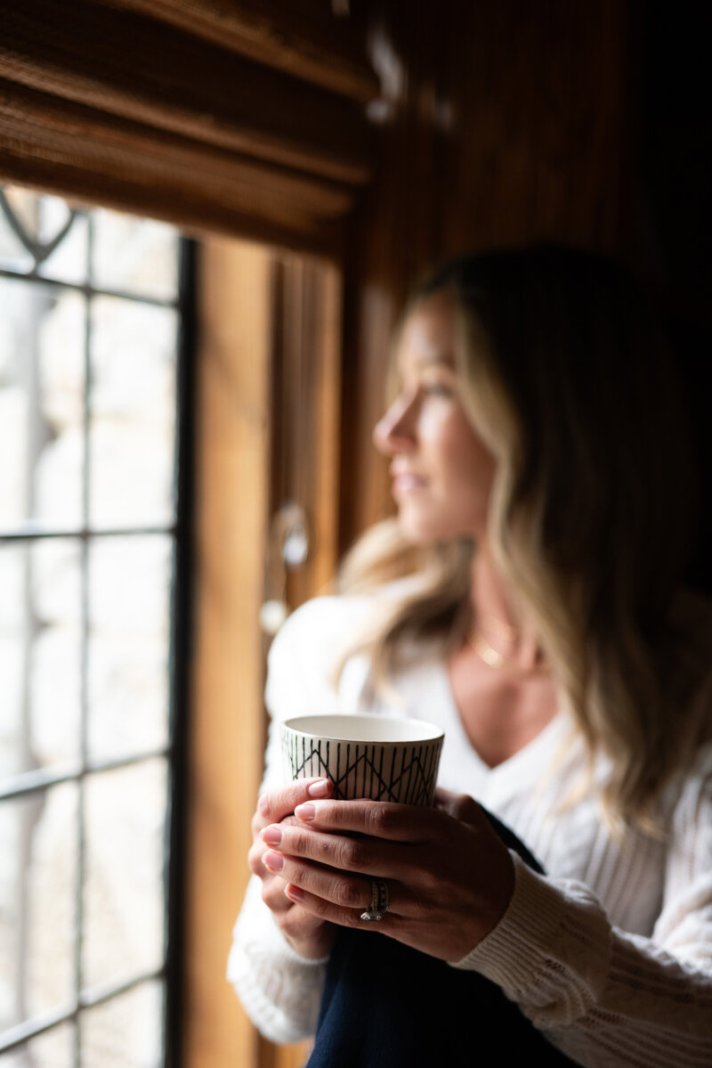 Jen, an integrative health coach, enjoying a cup of coffee at home while looking out the window, savoring a peaceful moment and reflecting on the day.