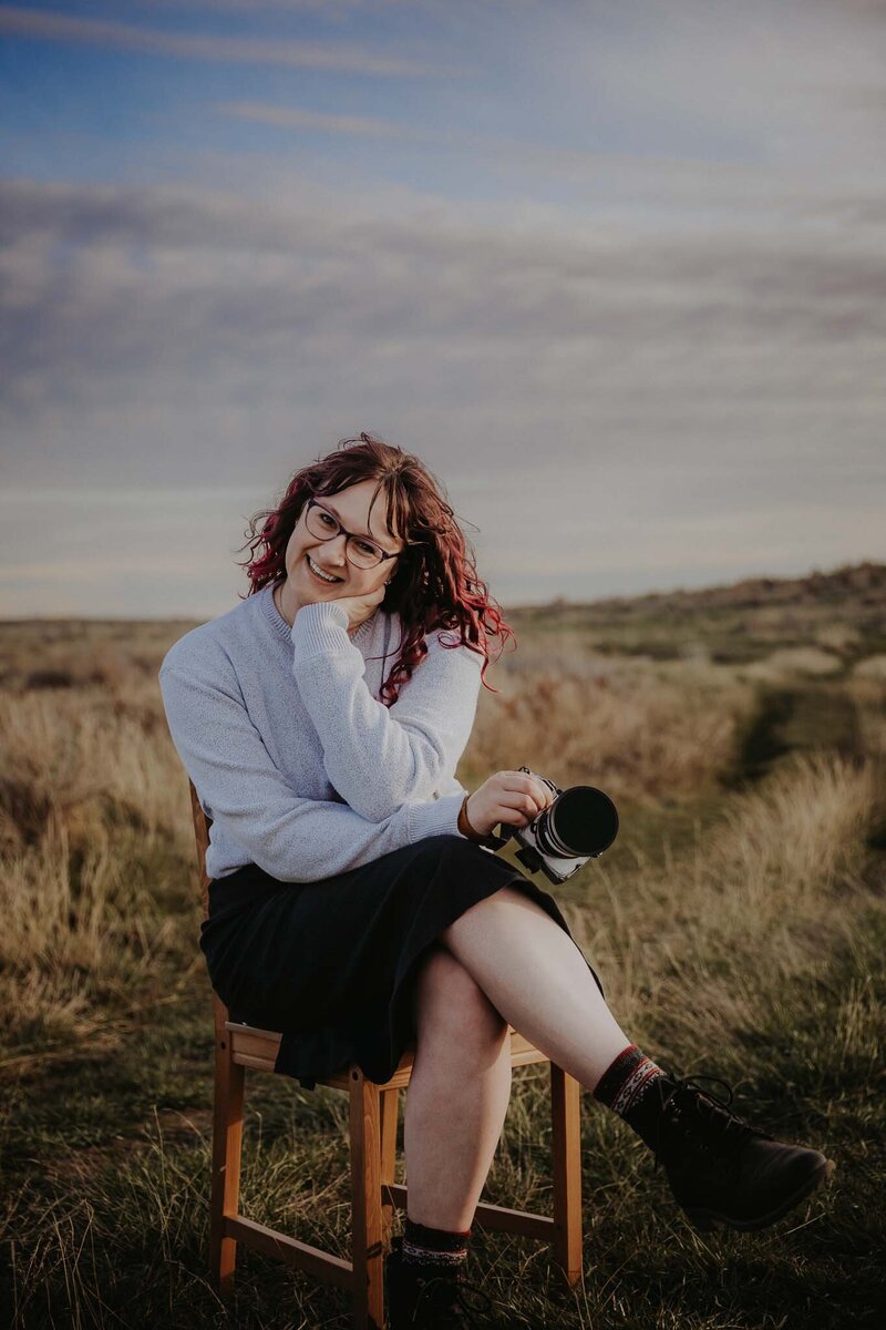 The photographer is crouched on a grassy hill with a lake behind her. She's holding a camera and looking to the right.