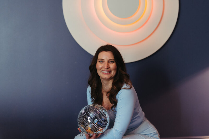 A woman in a light blue outfit beams while holding a disco ball, set against a dark blue wall with concentric circles—a vivid scene symbolizing creativity and joy in luxury event planning.