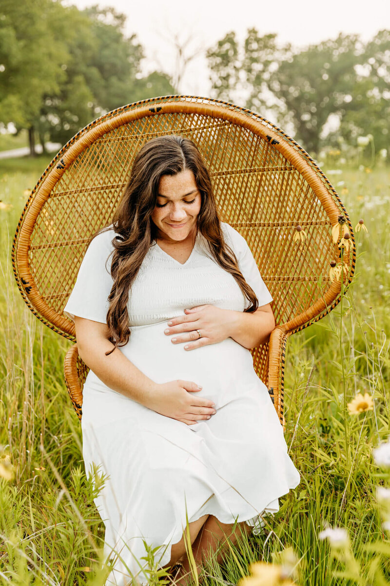 beautiful brunette pregnant woman smiling as she holds her bump while sitting in a peacock chair