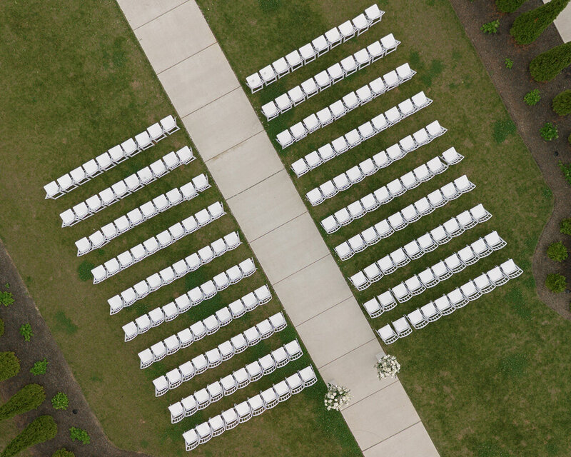 Rows of white chairs neatly arranged on grass flanking a central pathway, set for an outdoor event with floral arrangements at the path's end.