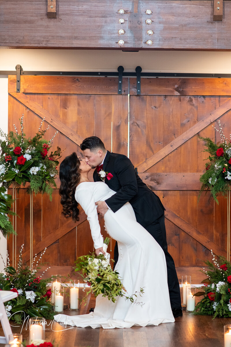 A couple shares a romantic kiss in front of large wooden double doors, capturing a classic barn wedding picture in Virginia. The bride holds a bouquet of white flowers, while the groom gently dips her amidst candlelight and floral arrangements at this enchanting outdoor wedding venue.