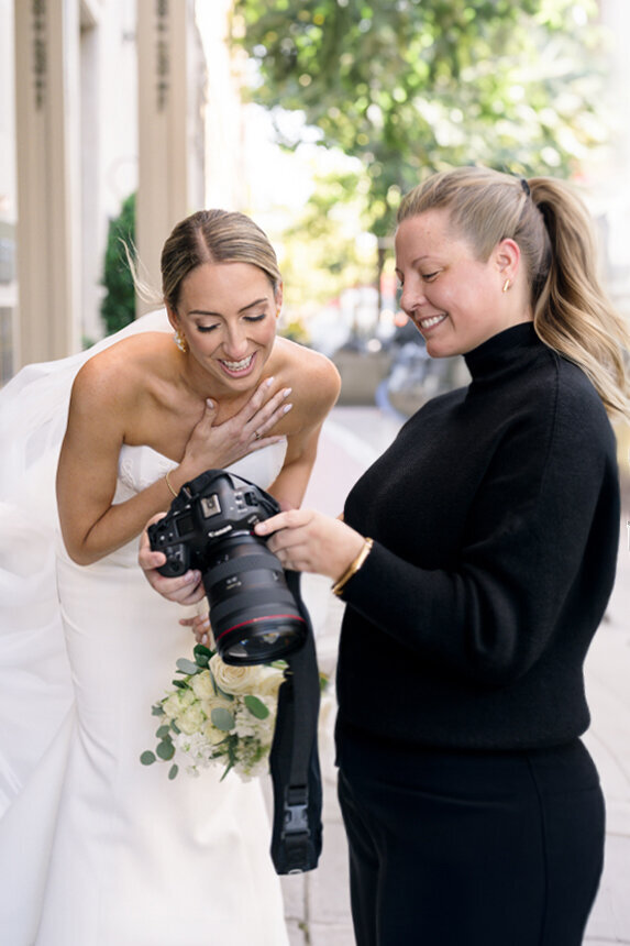 A bride in a white dress holds flowers and smiles while looking at images on a camera held by a woman in black. They are standing outside on a sunny day.