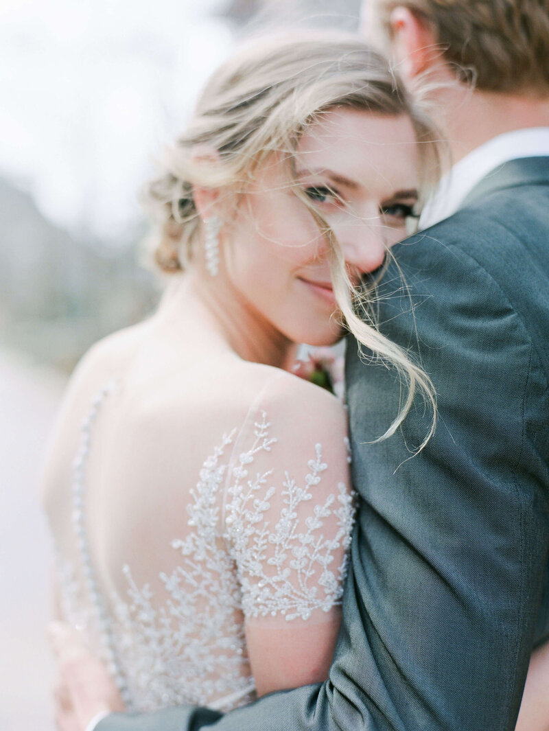 A bride, in the embrace of her groom, looks directly into the camera and smiles demurely, as the wind blows her blonde hair into her face.