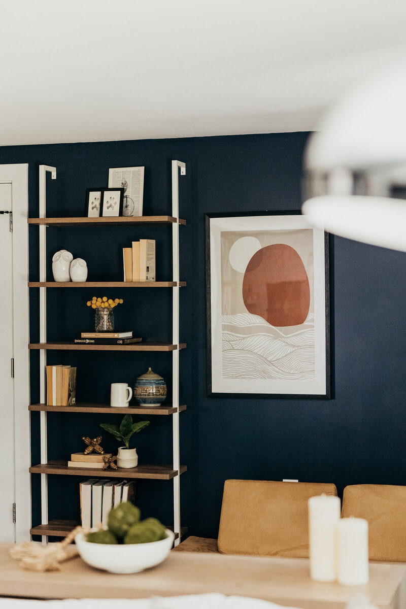 A close up of living room featuring a bookcase with various objects, a framed picture and seating area representing a colorful interior design by Winsome Home Interiors.