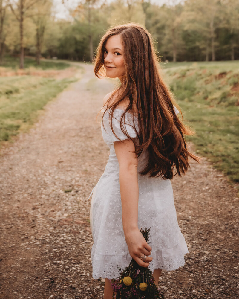 Beautiful senior girl in white dress holding flowers, walking down gravel road, looking back at camera