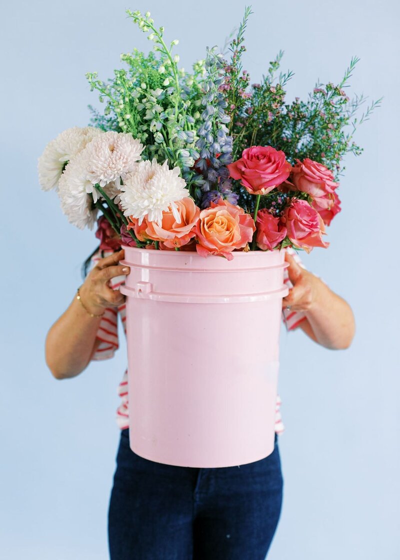 Pink bucket filled with vibrant garden roses, chrysanthemums, delphinium, and greenery held by florist in striped top