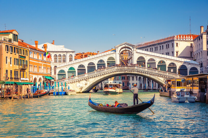 A gondolier navigates a gondola along the Grand Canal in Venice, Italy, under a sunny blue sky. The iconic Rialto Bridge, bustling with people, arches over the canal. Colorful historic buildings line the water's edge.