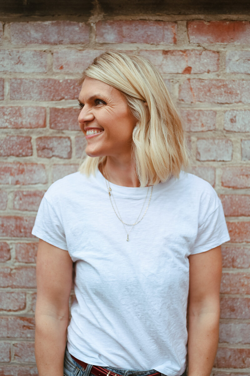 Emily Collins smiling in front of a brick wall, wearing a white tee and gold necklace.