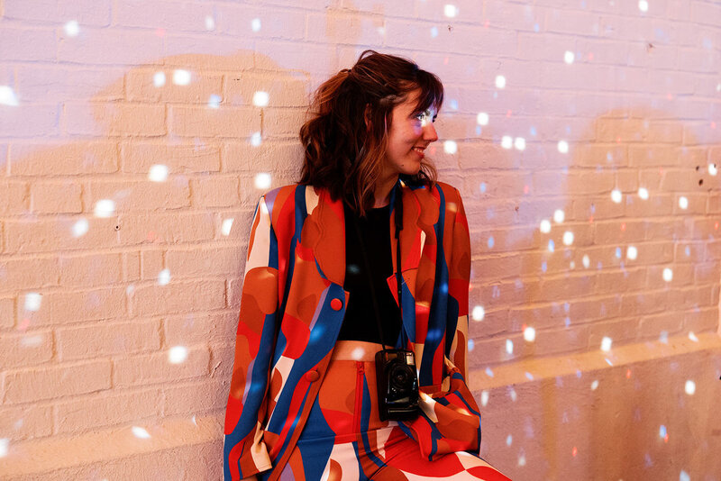 A film photographer holding a small analog film camera, wearing a funky suit of red orange blue white and purple, there are lots of little lights reflecting onto her and the white brick wall behind her. She is looking to the right smiling at someone dancing