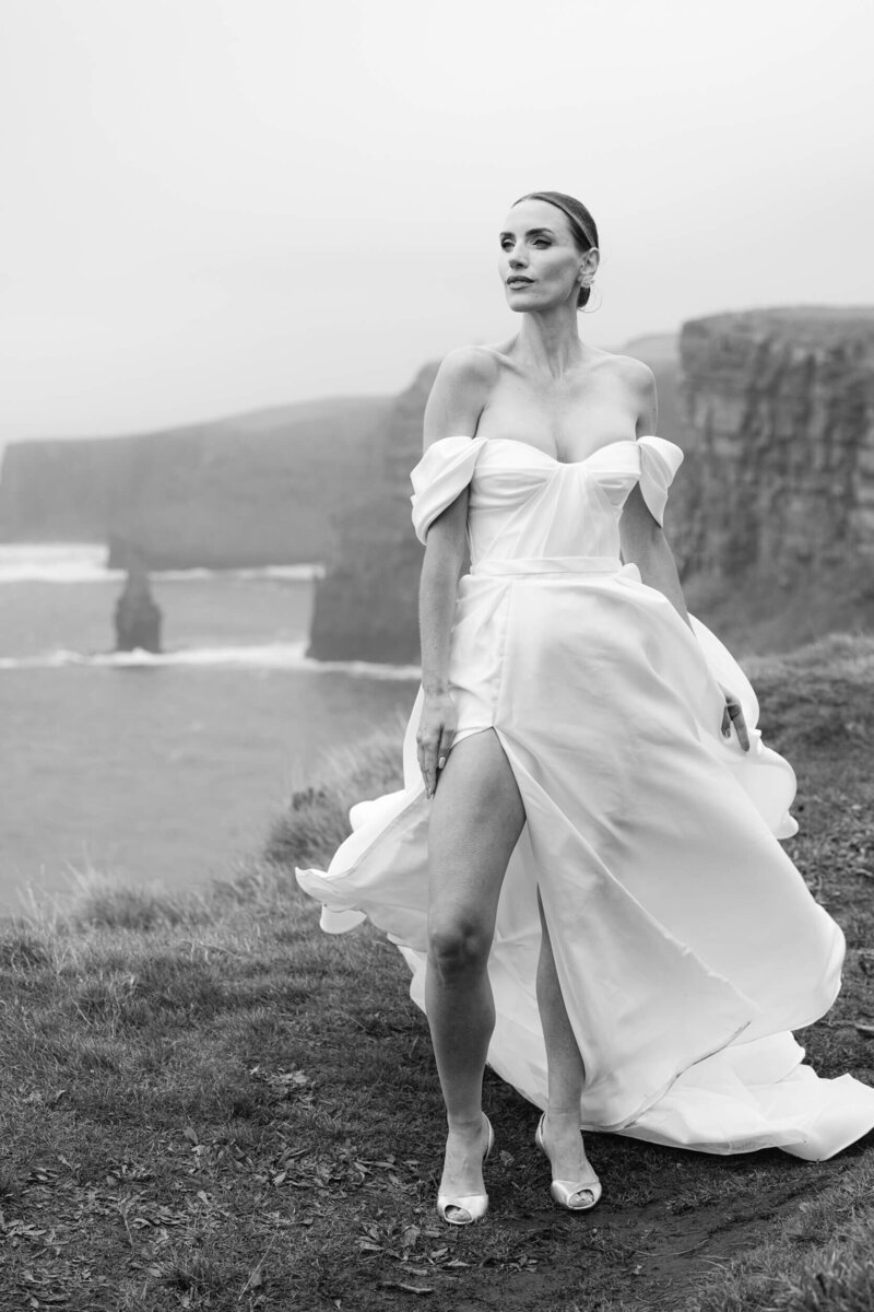 black and white image of a bride during her elopement standing at the top of the cliffs of moher with the ocean and cliffs in the background