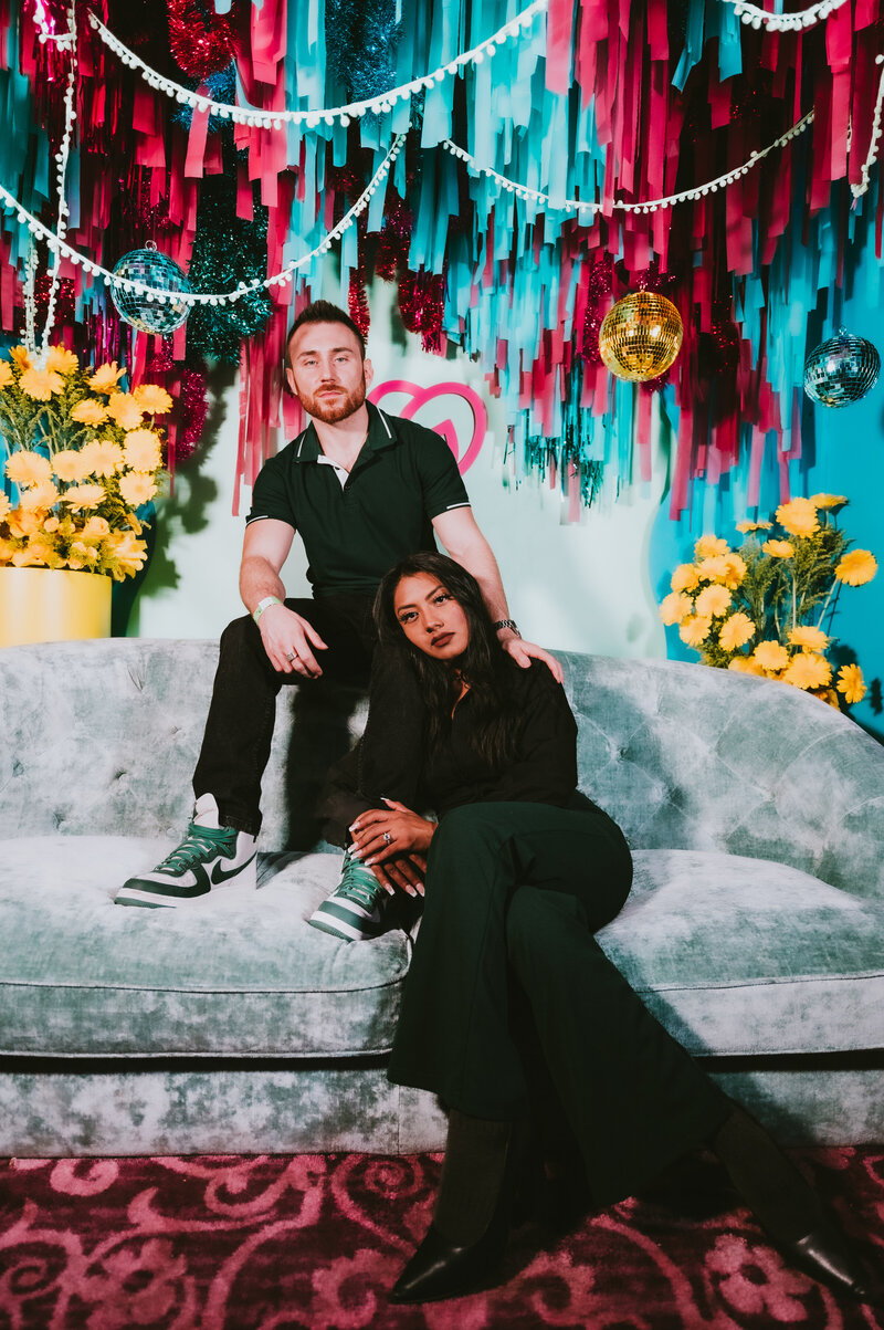 Two guests posing on a velvet couch in front of a vibrant, custom-designed photo booth backdrop with fringe, disco balls, and florals—captured by MISGIF at a branded corporate event in Phoenix, Arizona