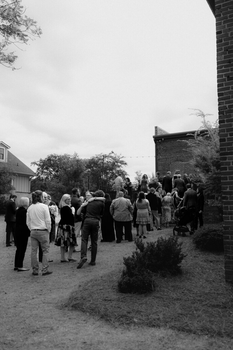 A group of wedding guests walk from the ceremony to the reception. A mother and son stop for an embrace as everyone moves around them.