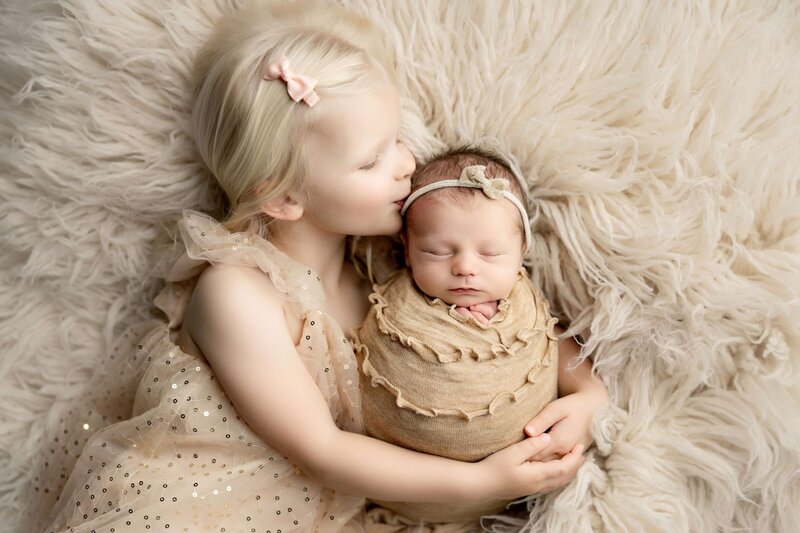 Big sister kisses her new baby sister on the forhead, snuggled together on a monochromatic cream and taupe fur