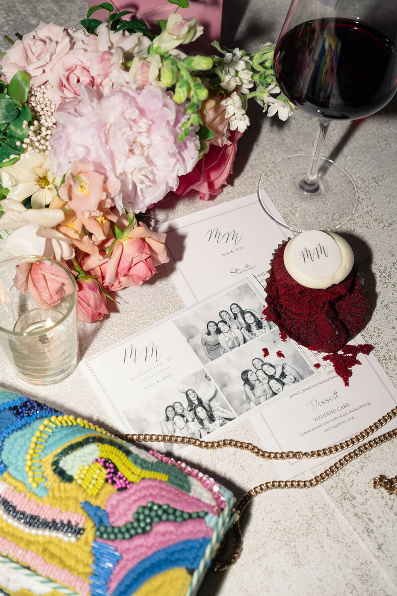 A table setting with a vibrant beaded purse, a glass of water, a glass of red wine, a red velvet cupcake, and event invitations featuring black-and-white photos. A bouquet of pink and white flowers adds elegance to the arrangement.