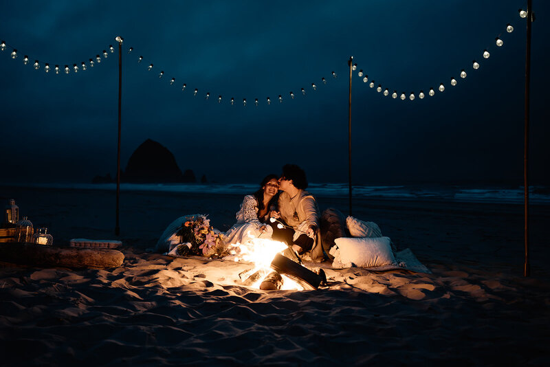 a couple snuggles in front of a bonfire in their wedding attire at the end of their adventure elopement package. The silhouette of Haystack rock can be seen behind them.   