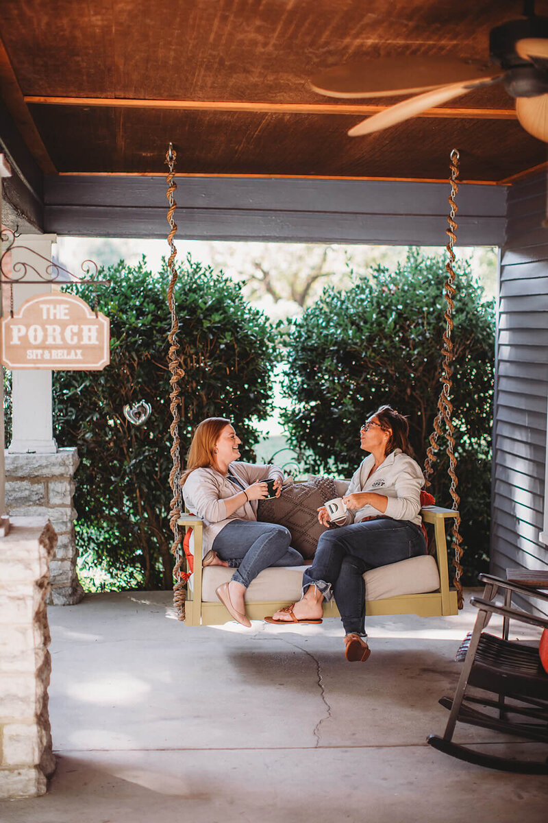 Two women sitting on a porch swing at Matters of the Heart Counseling Center, engaged in conversation. They're holding mugs and appear relaxed. A sign reading "THE PORCH - SIT & RELAX" is visible. The porch has a wooden ceiling with a fan, and is surrounded by greenery, creating a peaceful atmosphere.