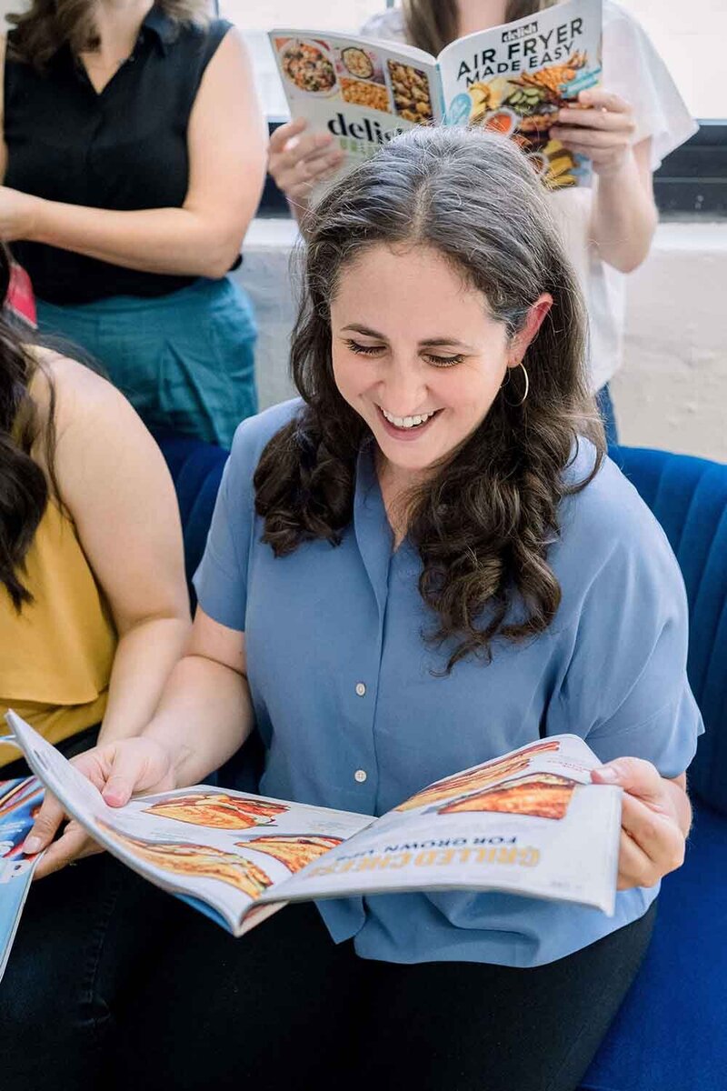 Woman sitting in a chair wearing a blue shirt looking at a food magazine and smiling.