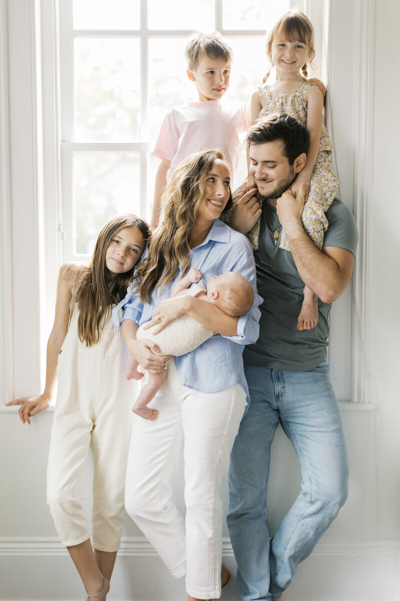 A joyful family of five gathered by a bright window, smiling and snuggling their newborn during an in-home session — Raleigh portrait photography.