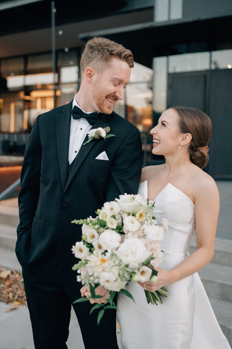 Groom in black tux and bride in asymmetrical wedding dress with a v neck smile at each other in front of The Farnam Hotel in Nebraska. Photo by Anna Brace, an NE Wedding Photographers.