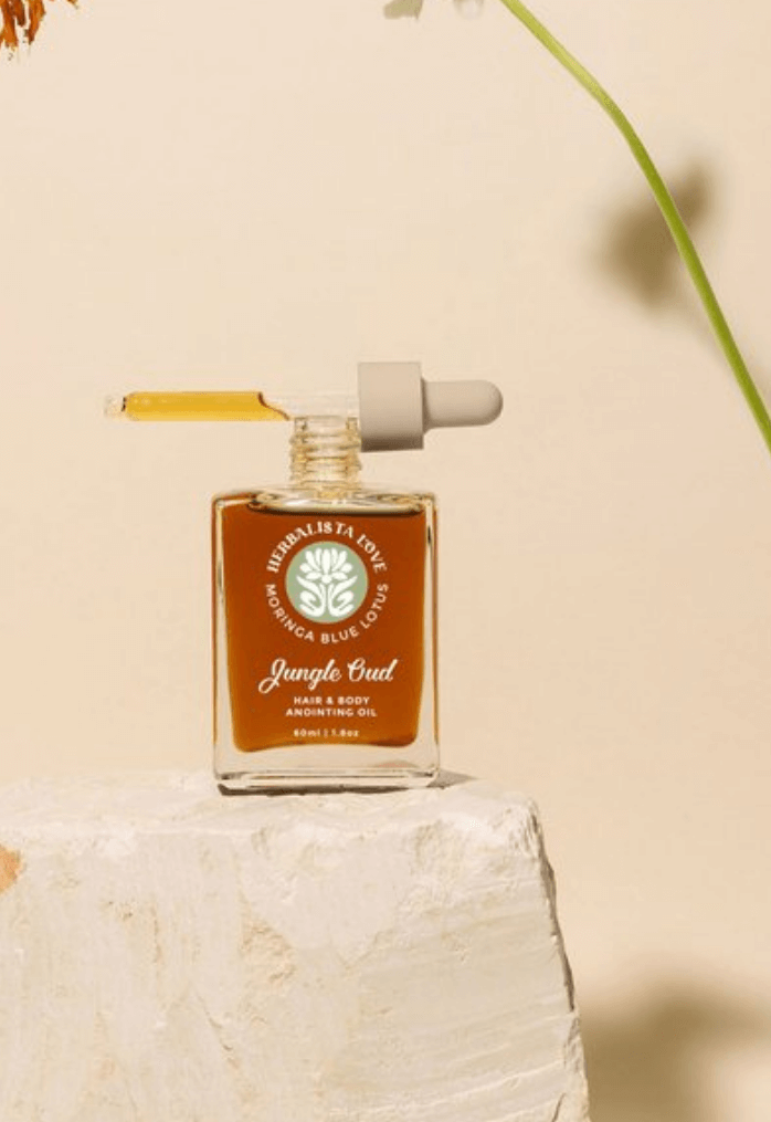 A square amber bottle of Herbalista Love Jungle Mud facial oil, displayed on a textured stone surface against a minimal background. The design emphasizes the product’s earthy and organic ingredients.
