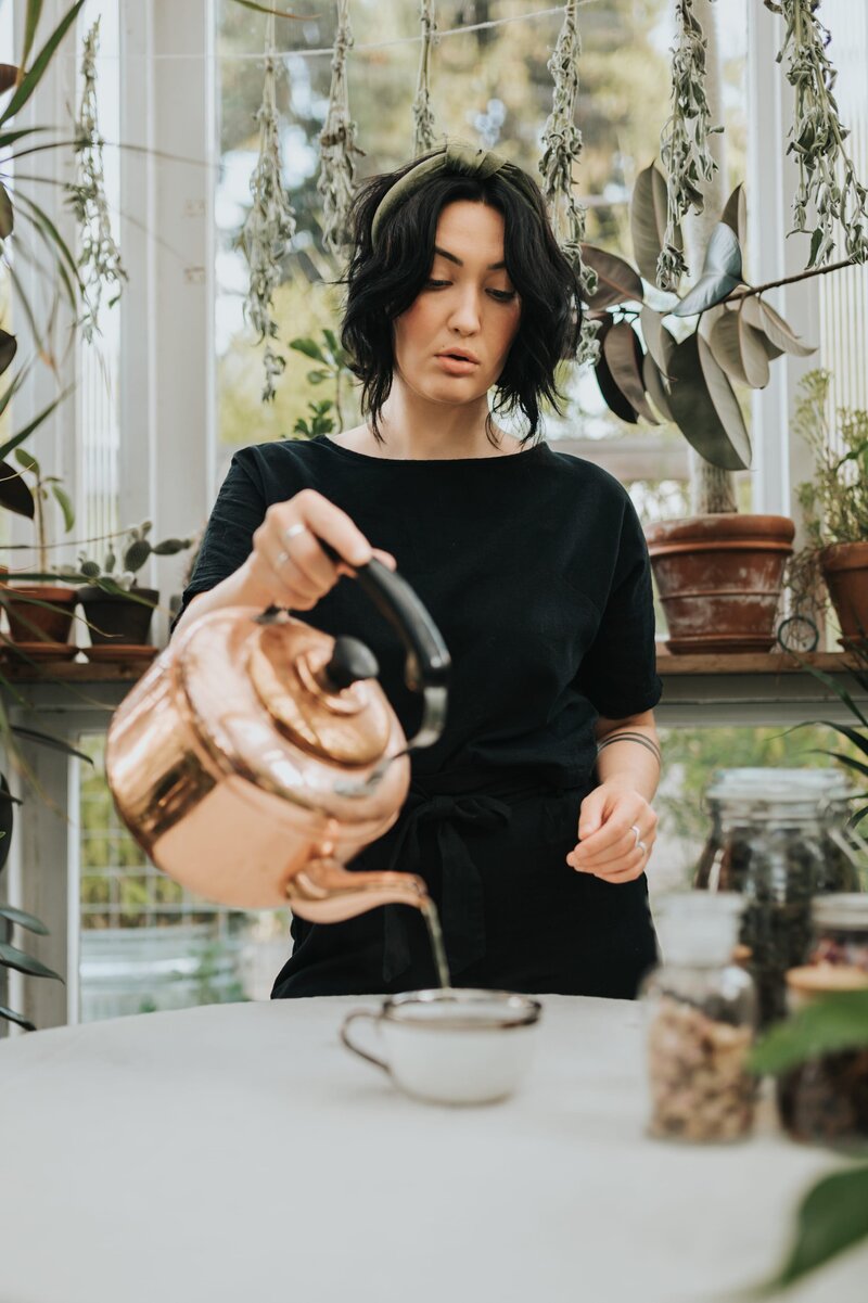 Woman wearing black shirt pours hot water from a copper kettle into a teacup surrounded by plants in a greenhouse.