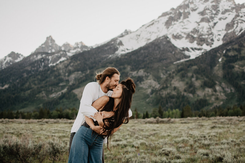 jackson hole photographers captures engaged couple standing in the Tetons, the man is behind the woman and holding her in his arms while kissing her forehead