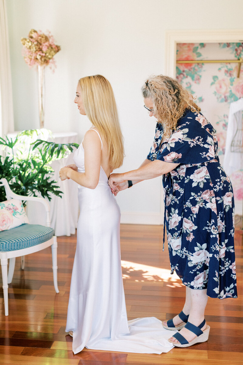 Bride is getting ready while mom helps her button her dress.