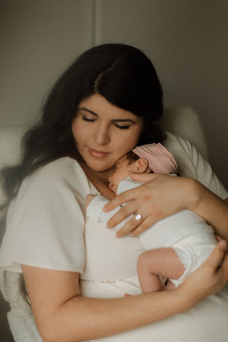 Mother gently holding her newborn daughter, wrapped in white with a soft pink bow, during a serene in-home newborn session in Canyon Lake, California.