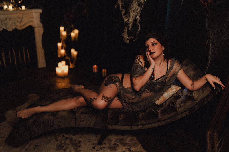 Woman with dark lipstick and sheer robe reclines on a chaise surrounded by candlelight and cobwebs for a dramatic spooky-season boudoir look