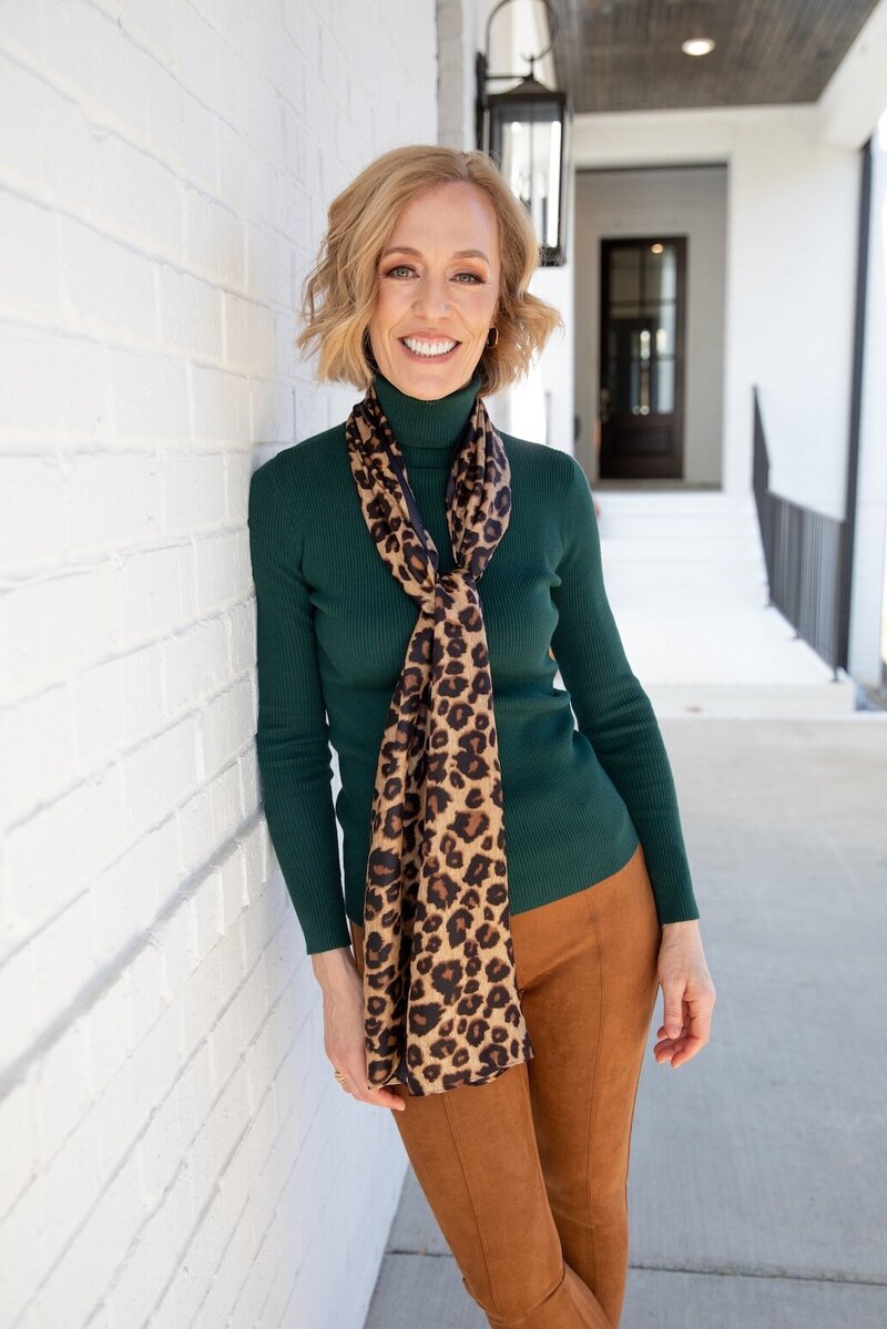 Heather Ebert stands outside a beautiful home wearing a rust-colored shirt dress. Her arms are crossed, and she's leaning against a white wall, smiling.