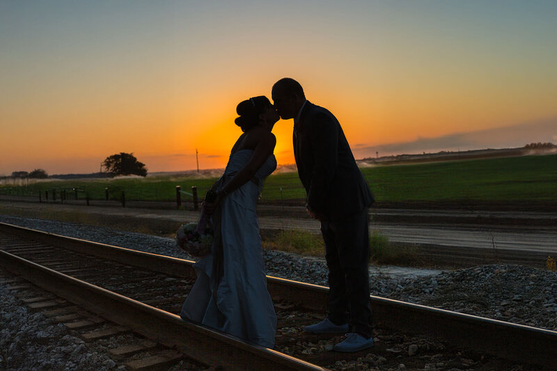 Bride and groom share a romantic silhouette kiss on the train tracks at sunset after their wedding ceremony.