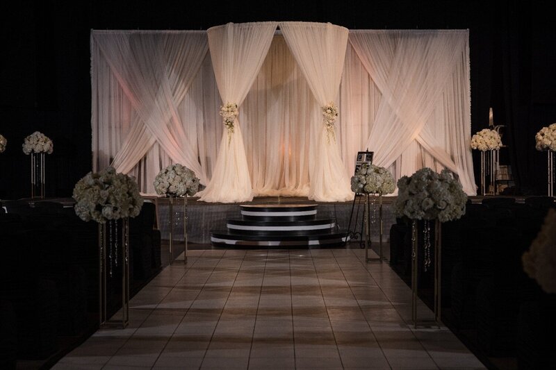 Elegant wedding ceremony altar setup at The Crane Bay Event Center in Indianapolis, featuring sheer white drapery, floral arrangements of white hydrangeas and roses, and a raised platform with modern black-and-white steps.