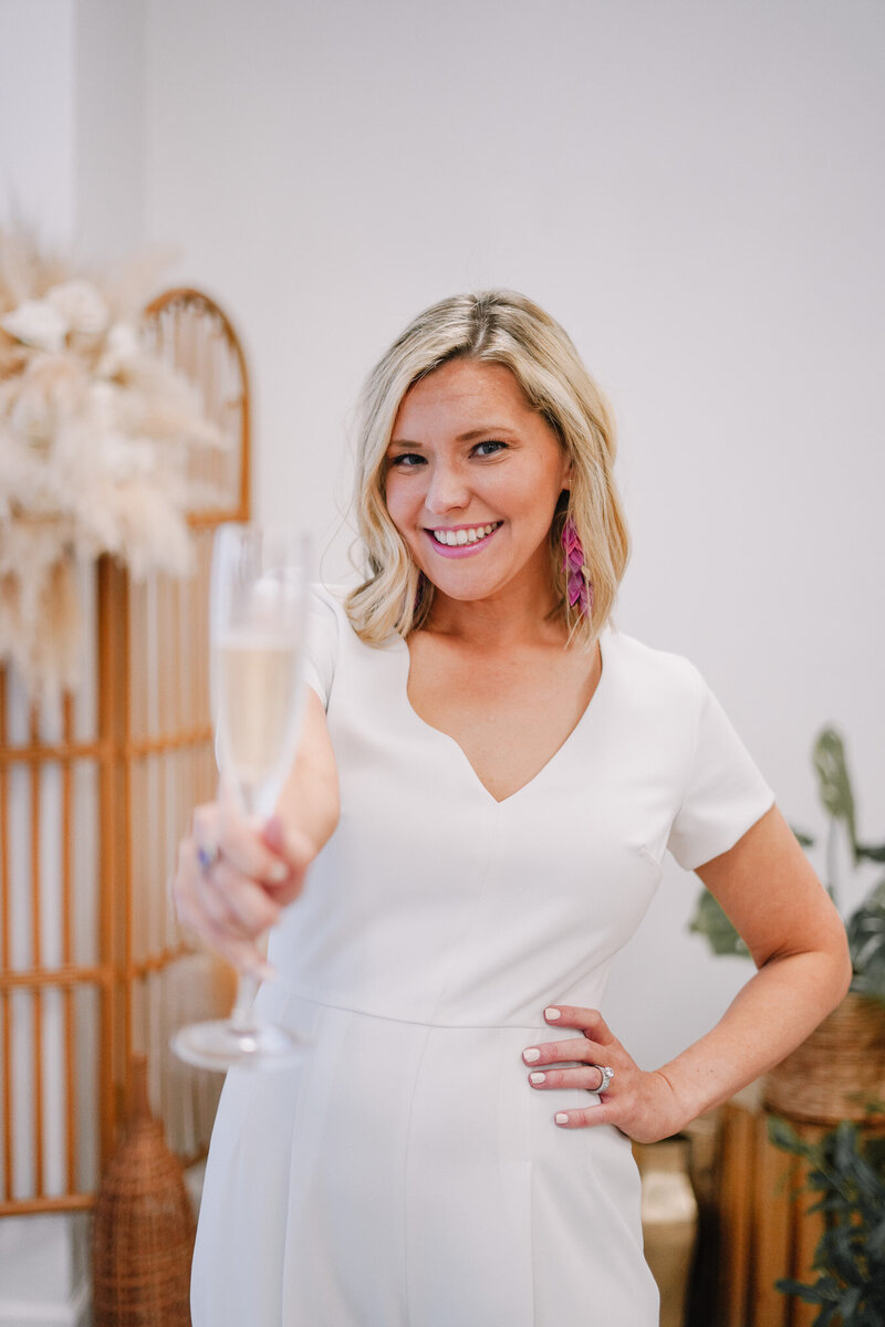 Woman wearing white dress smiles in creative studio while holding champagne flute up to camera