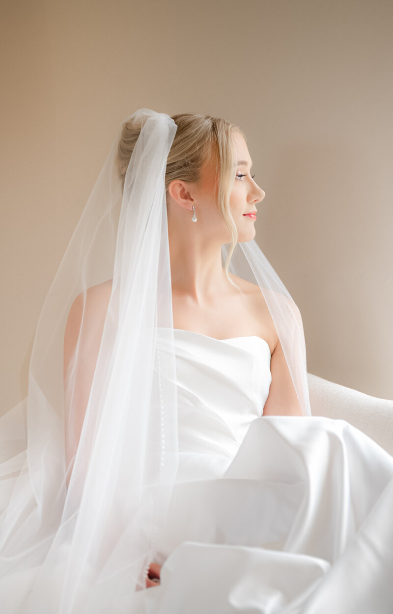 An editorial portrait of a bride with a long veil looking out the window into natural light