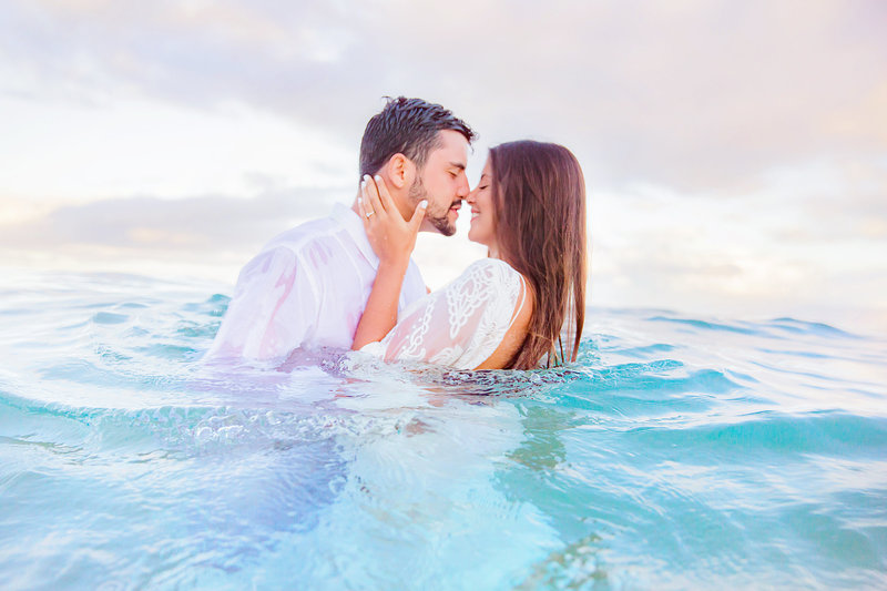 couple wearing white embraces in clear blue water leaning in for a kiss