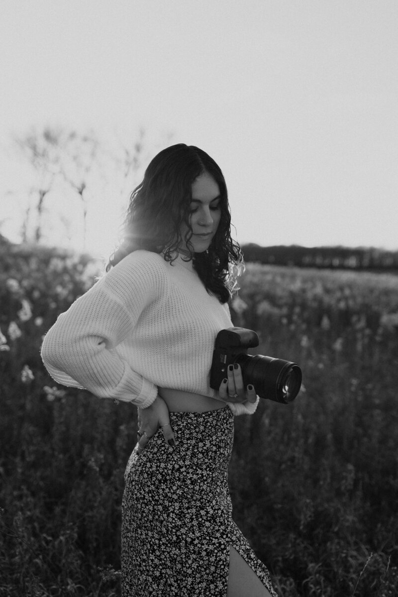 Candid black-and-white portrait of a female photographer holding her camera in a sunlit field, highlighting her creative spirit and connection with nature