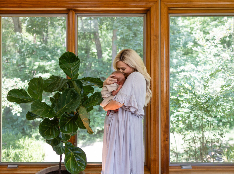 A mother in front of wooden window frames with a view and a green plant holding her infant son taken.