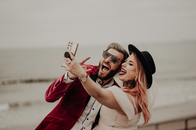 bride holding phone to take a selfie of her and groom, sticking toungues out bride wears black hate and groom wears red blazer and sunglasses