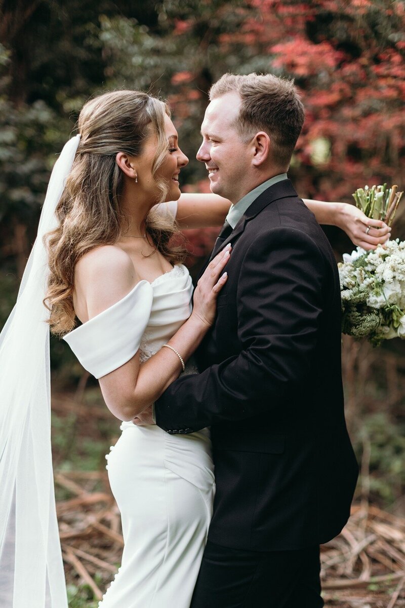 A bride wearing a white dress and veil and holding a bouquet, hugging a groom in a black suit.