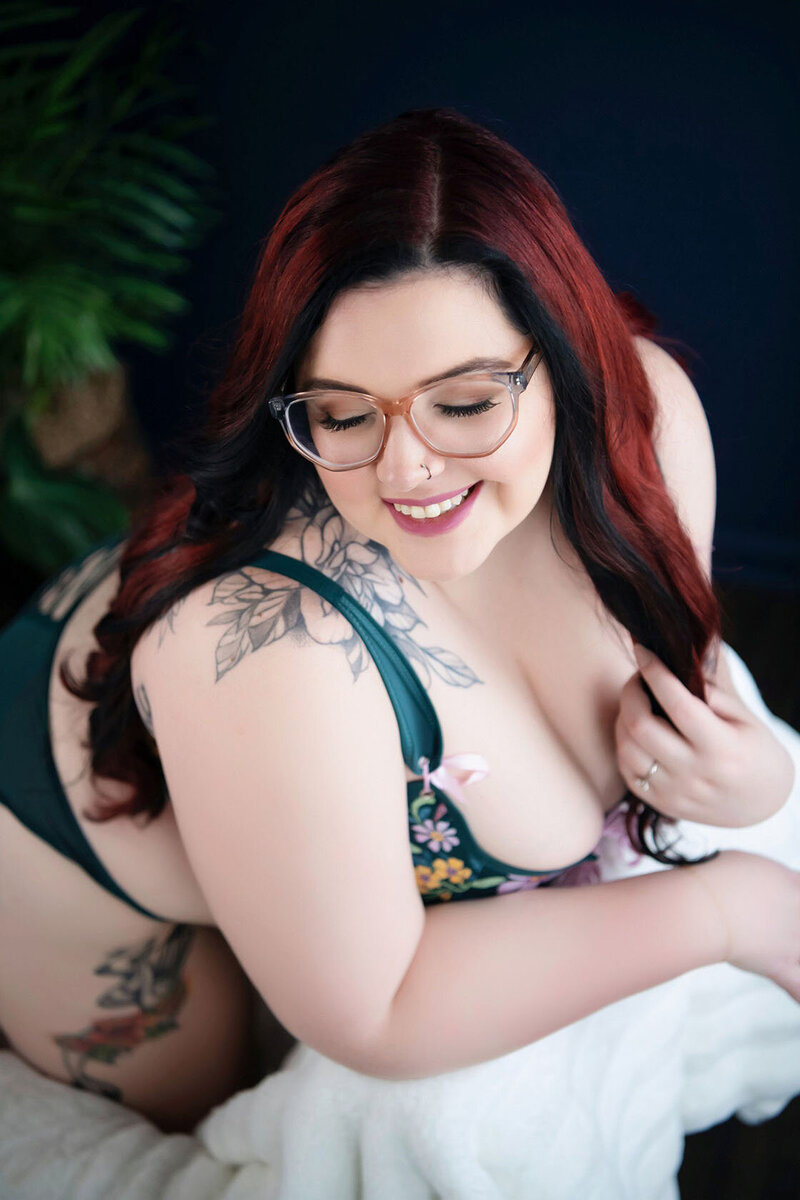Smiling woman with long dark hair and glasses, wearing a floral outfit. She shows a tattoo on her shoulder, exuding confidence and joy.