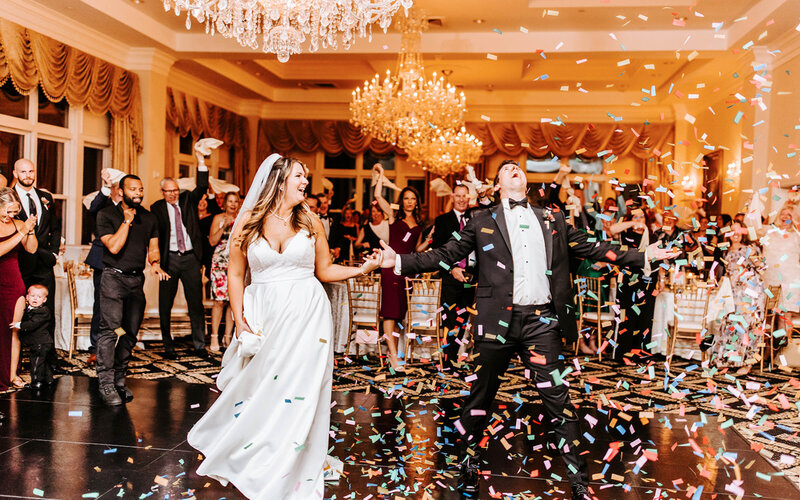 Joyful Bride and Groom, holding hands after entering wedding reception in the midst of confetti flying everywhere, as guests cheer in the back ground.