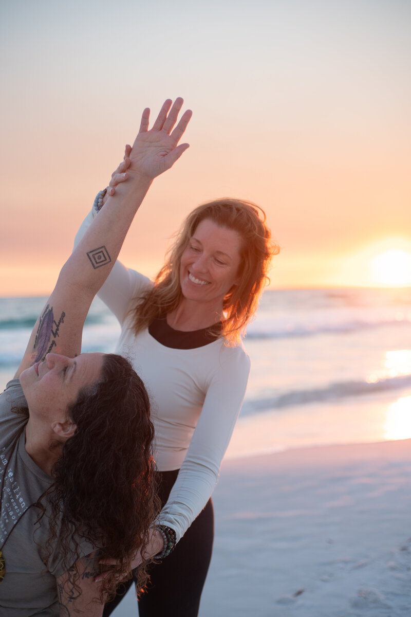 Stephanie assisting a woman in a yoga pose on the beach 