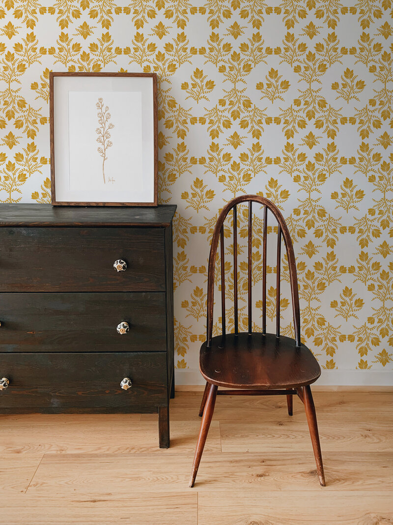 vintage hand drawn flowers in a symmetrical print in the color of sunshine or mustard yellow on a white background. Room has vintage wooden dark stained furniture, wallpapered walls, and a simple botanical art print